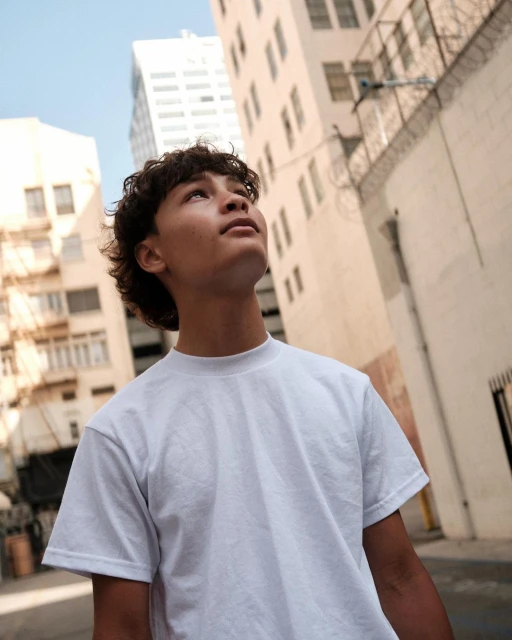 Low-angle shot of a young man in a white t-shirt looking up in an urban setting, with tall buildings visible in the background.