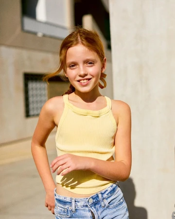 Young girl with red hair wearing a pale yellow tank top and denim shorts outdoors in sunlight