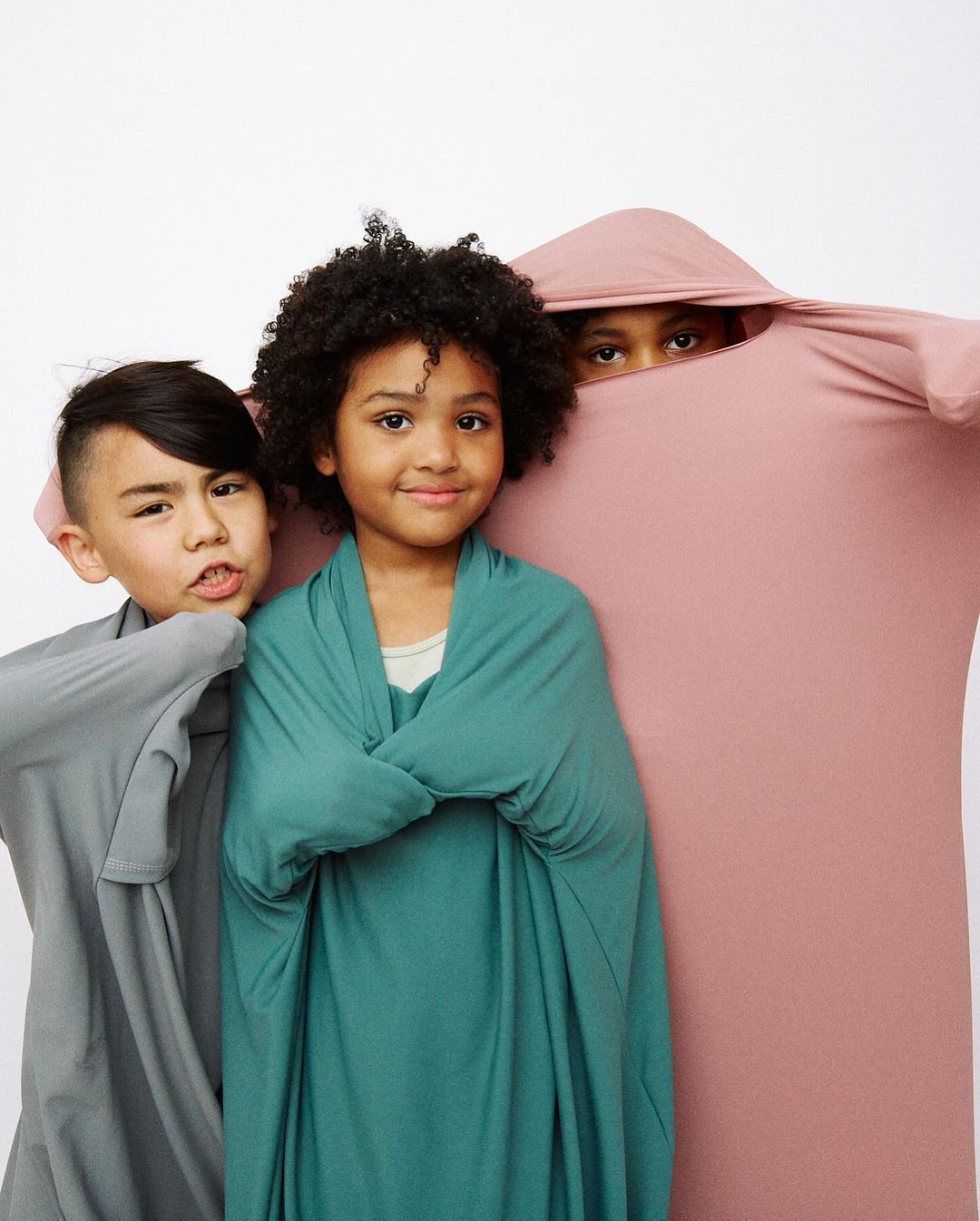 Three children playfully wrapped in colorful fabric against a white backdrop, creating a cozy and cheerful portrait, showcasing fabric textures.