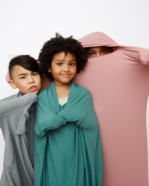 Three children playfully wrapped in colorful fabric against a white backdrop, creating a cozy and cheerful portrait, showcasing fabric textures.