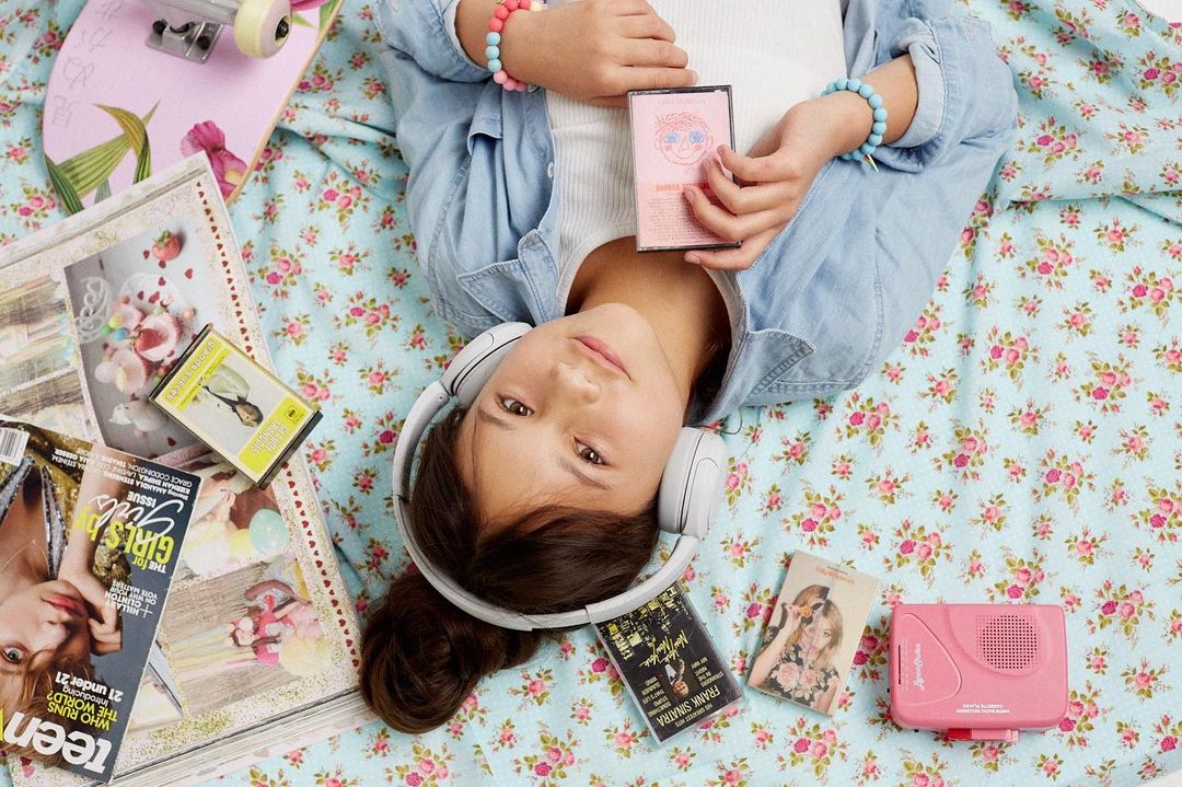 A young person with headphones lying on a floral blanket surrounded by music cassette tapes, a skateboard, and fashion magazines.
