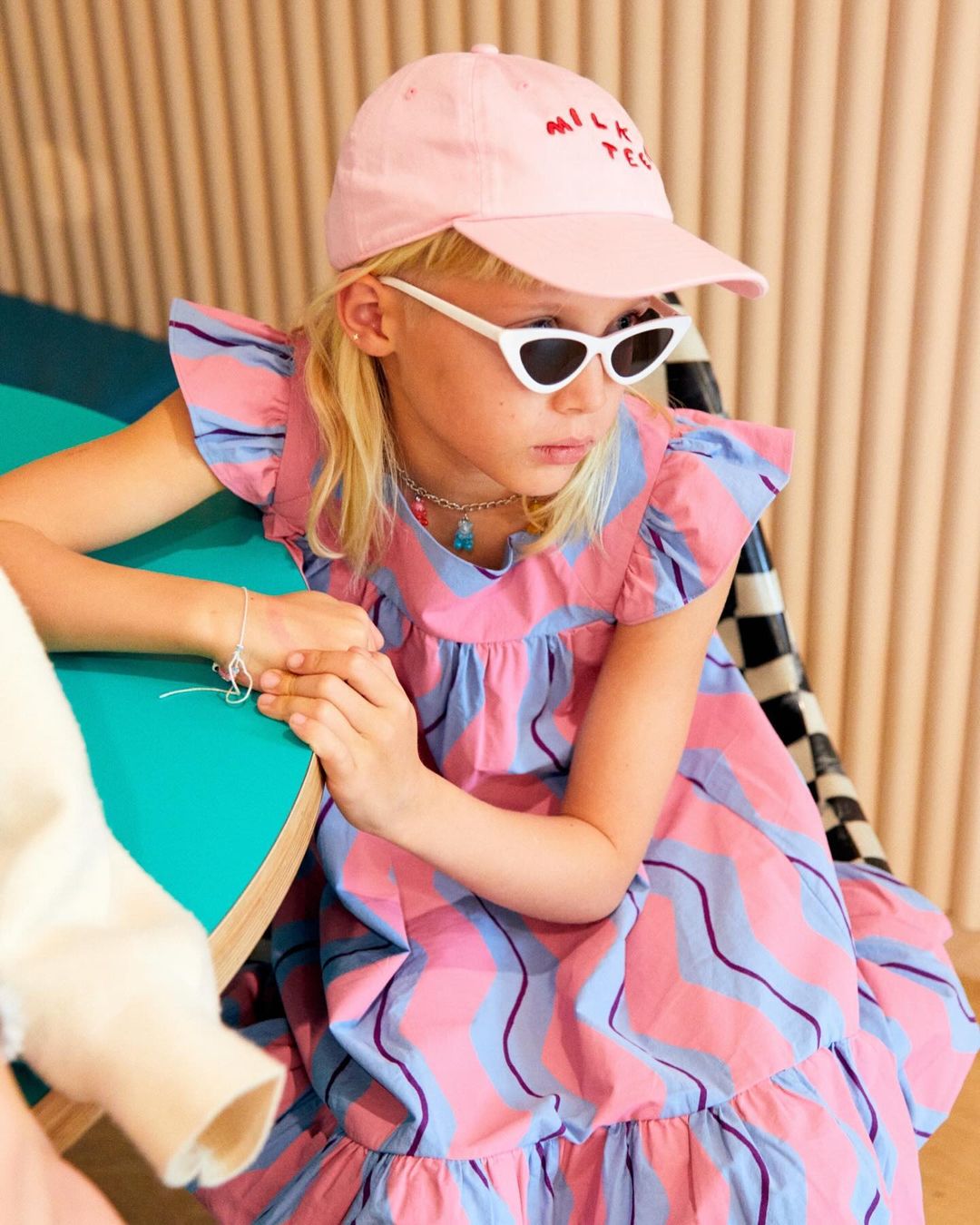 Young girl in pink cap and striped dress, wearing sunglasses, poses indoors near a turquoise table and checkered chair.