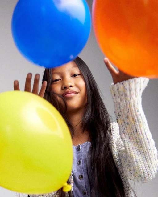 Young girl smiles surrounded by three colorful balloons against a light grey backdrop in a studio setting, creating a joyful, vibrant atmosphere.