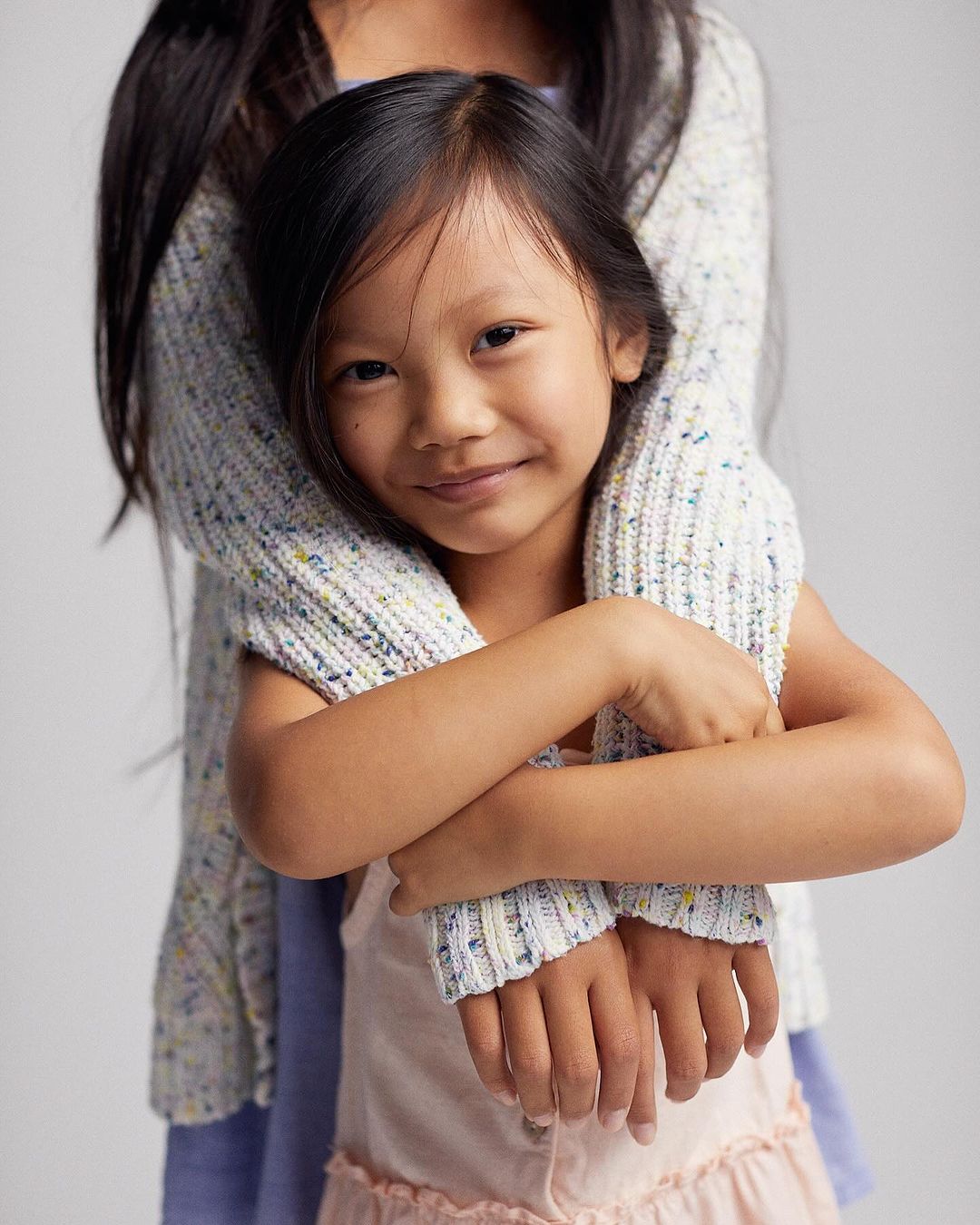 A young girl with dark hair smiles at the camera while being embraced from behind by an adult wearing a textured knit sweater.