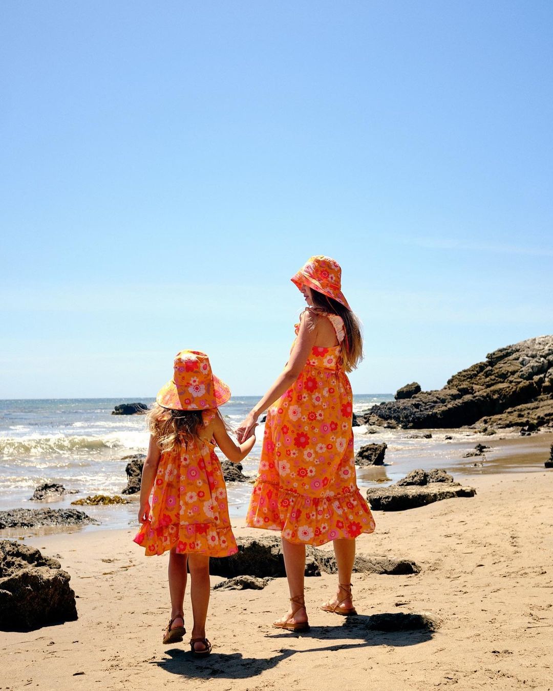 A woman and a child in matching orange floral dresses and hats walk hand-in-hand on a sandy beach toward the ocean on a sunny day.