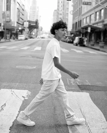 Young man walking across a city street intersection wearing a plain t-shirt and trousers in a monochrome photograph.