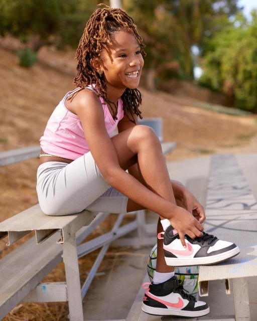 Smiling girl with locs sits on a metal park bench outdoors tying a black-and-white sneaker with a pink swoosh at daytime.