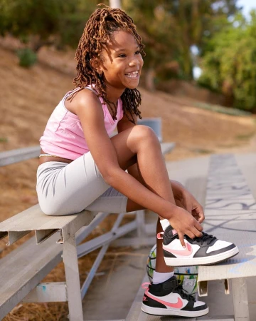 Smiling girl with locs sits on a metal park bench outdoors tying a black-and-white sneaker with a pink swoosh at daytime.