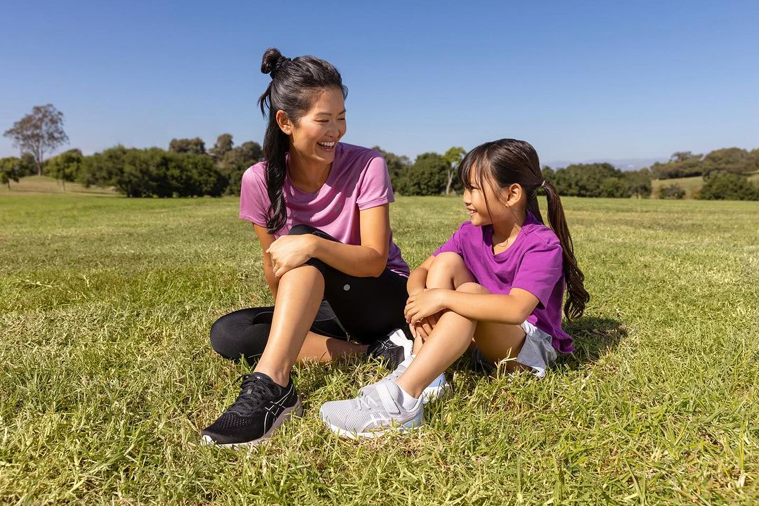A happy adult woman and a child sit on a grassy field, smiling at each other on a sunny day, both wearing athletic clothing and sneakers.