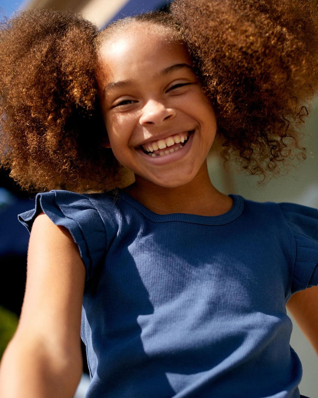 Close-up of a young girl with large, curly hair smiling broadly, wearing a blue ribbed top. Outdoor setting with natural sunlight.