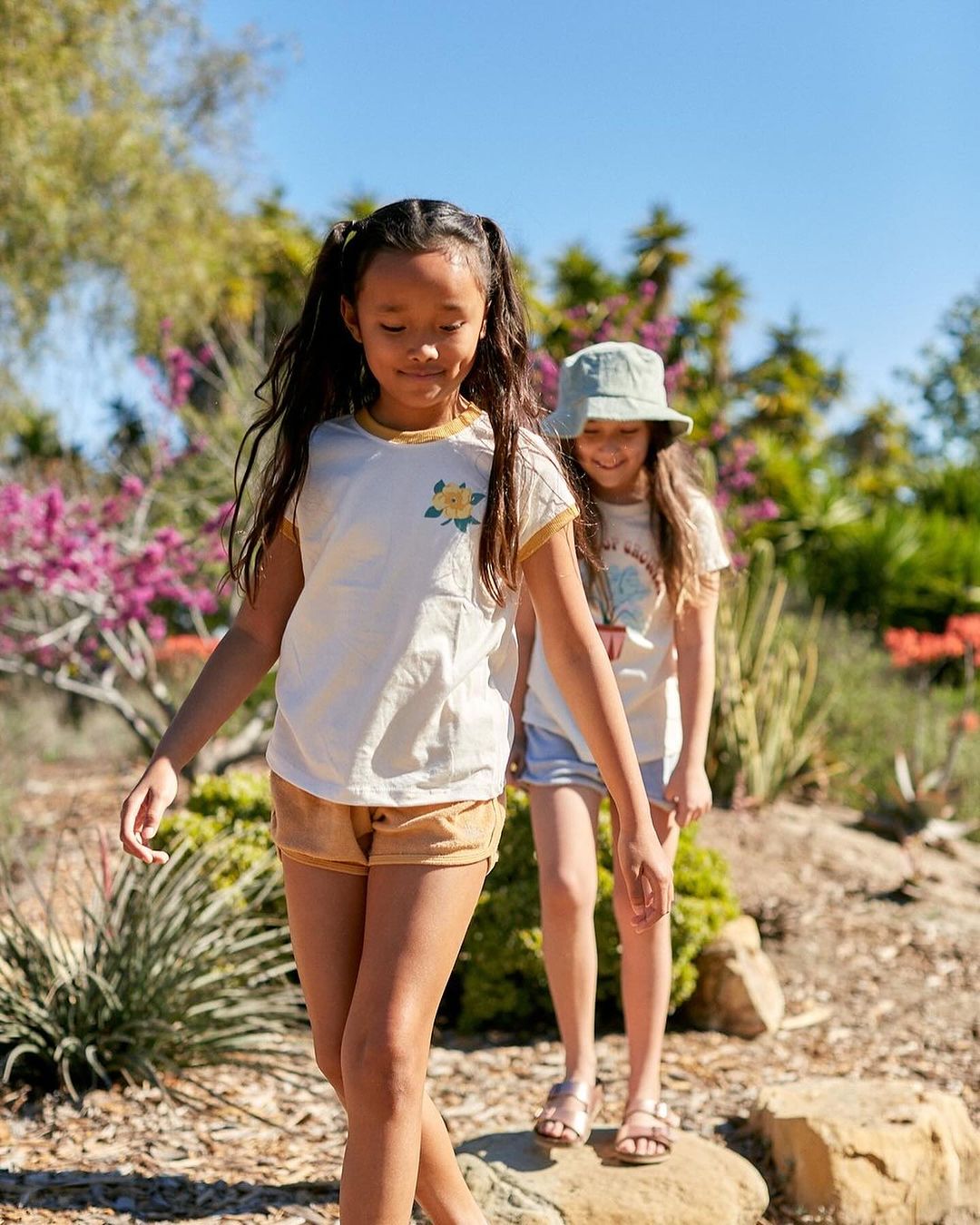Two girls in casual clothing walk on a stone path in a sunlit outdoor setting with vibrant flowers and lush greenery in the background.
