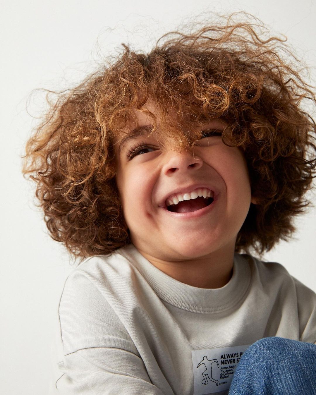 Portrait of a laughing child with curly brown hair wearing a light t-shirt against a plain white background