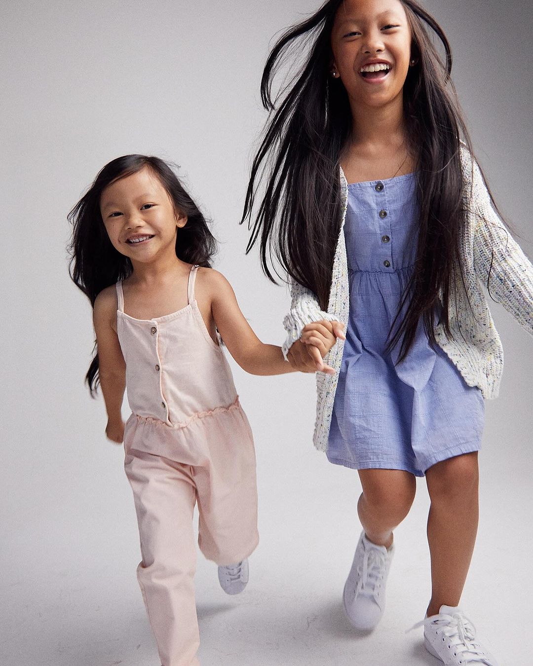 Two happy young girls running indoors, holding hands, and laughing. One wears a blue dress and the other a pink jumpsuit.