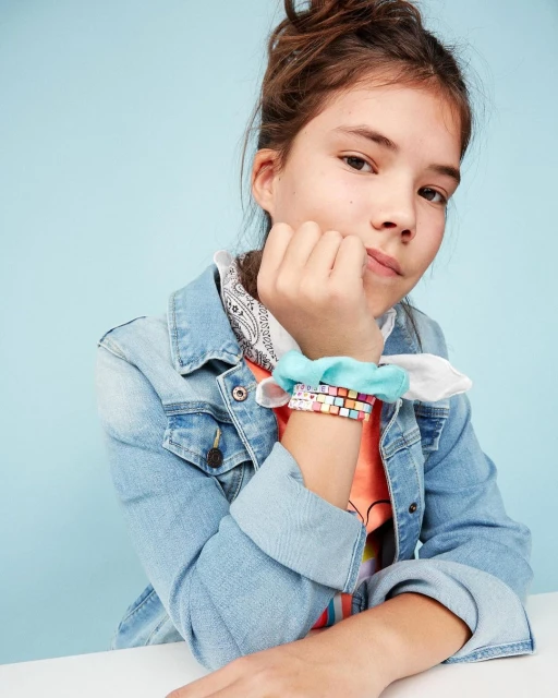 Portrait of a young girl with brown hair in a bun, wearing a denim jacket and colorful bracelets, sitting at a white table against a blue backdrop.