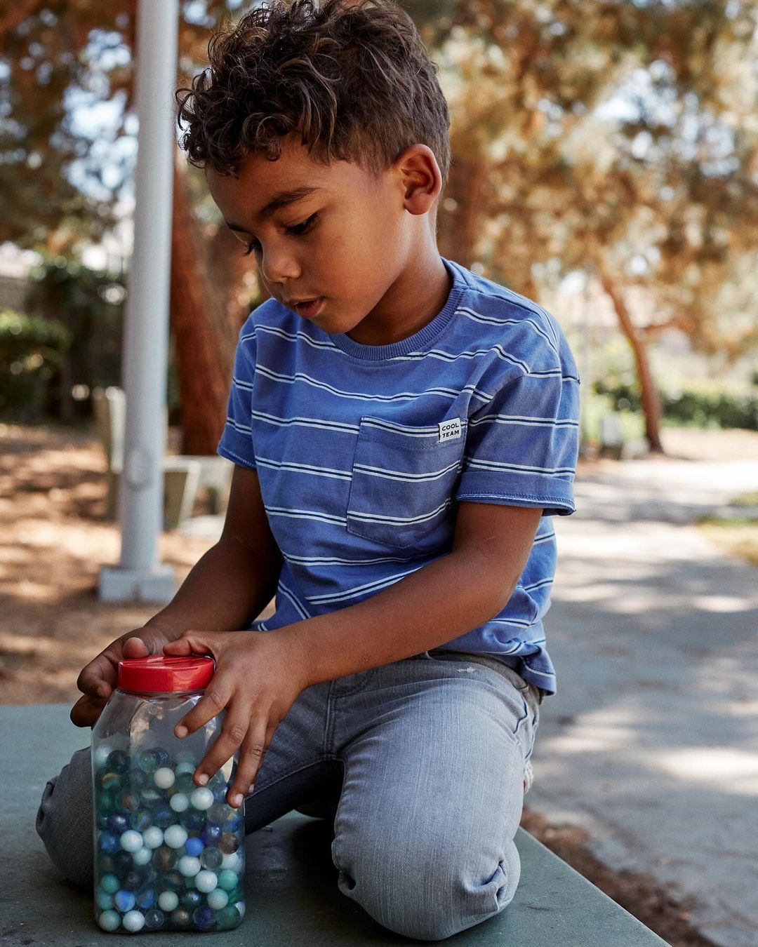 A young boy sits outside wearing a blue striped t-shirt, looking at a jar filled with marbles, with a background of trees and a park.