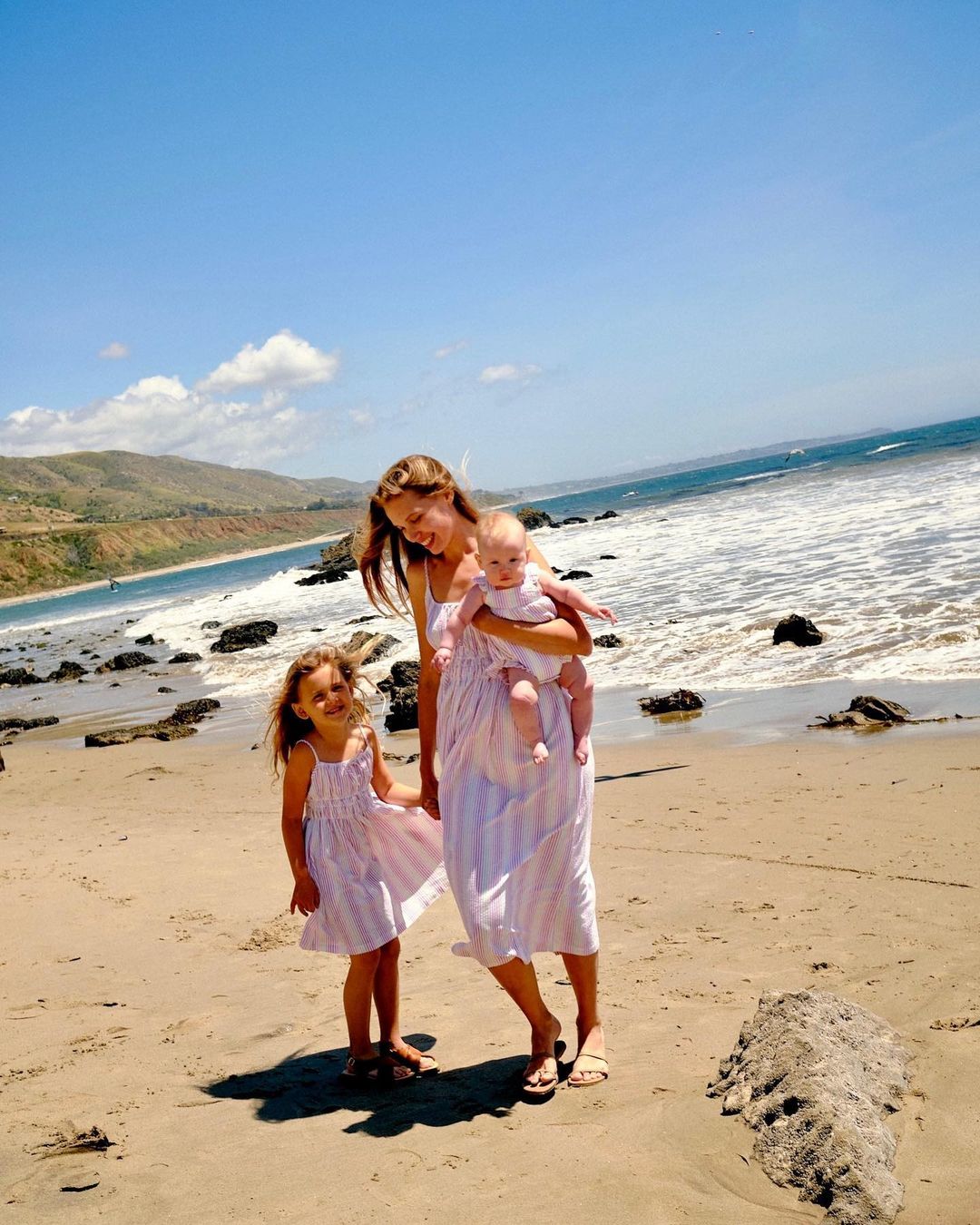 Mother and two daughters in matching dresses on a sandy beach with waves and a coastal landscape in the background under a blue sky.