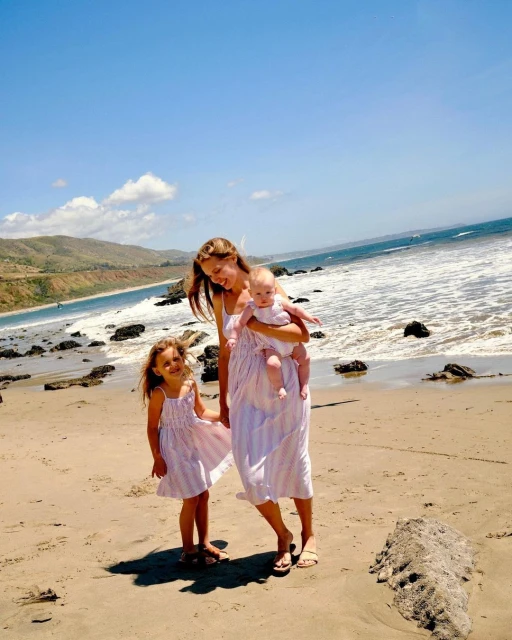 Mother and two daughters in matching dresses on a sandy beach with waves and a coastal landscape in the background under a blue sky.