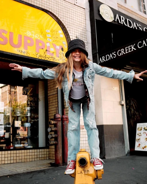 A young girl with a bucket hat stands on a yellow fire hydrant in an urban setting, smiling with arms outstretched, wearing a denim jacket.