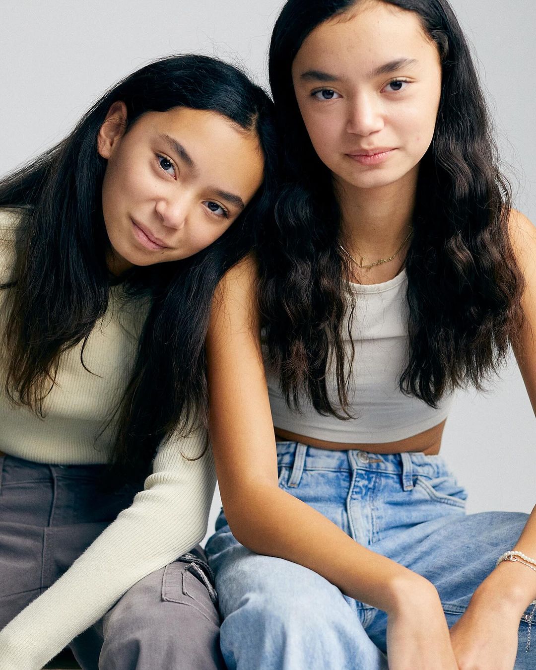 Two young women pose indoors in casual clothing. One rests her head on the other's shoulder in a portrait-style fashion shot.