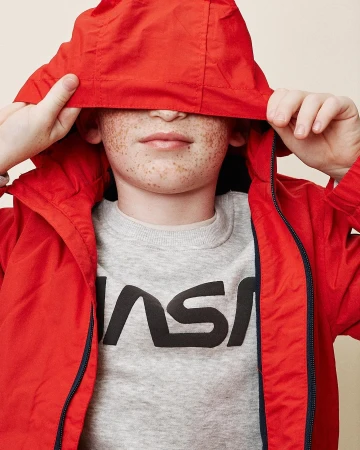 Young child wearing a red hooded jacket and grey NASA sweatshirt indoors in a close-up shot, showing freckles and red hood.