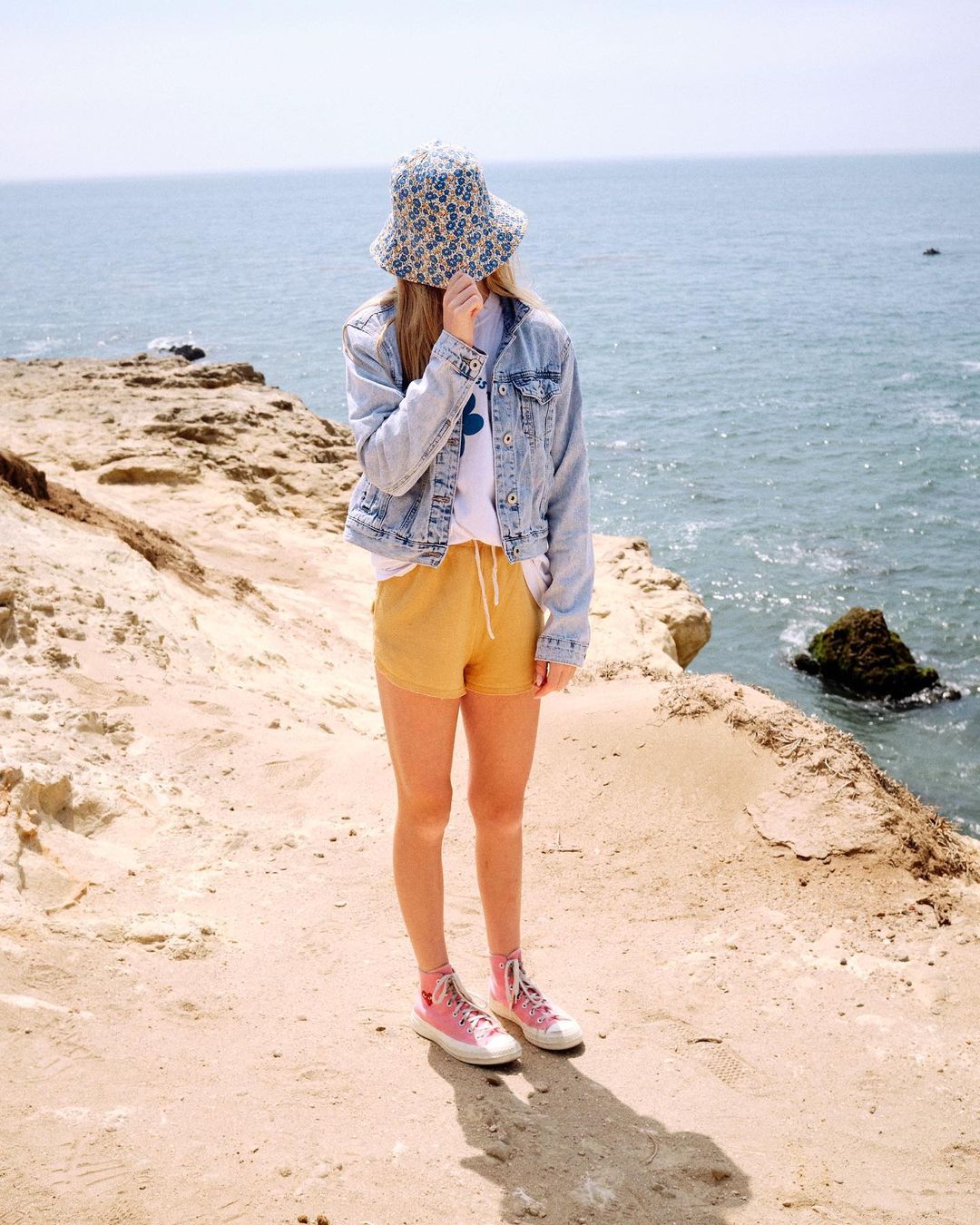 Young woman wearing a patterned bucket hat, denim jacket, yellow shorts, and pink sneakers, stands on a sandy path by the ocean.