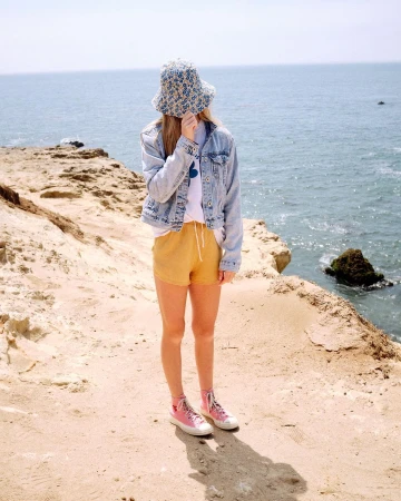 Young woman wearing a patterned bucket hat, denim jacket, yellow shorts, and pink sneakers, stands on a sandy path by the ocean.