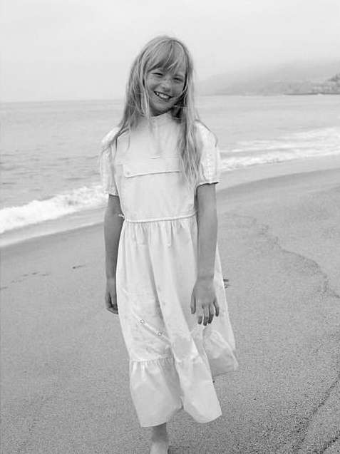 A young girl with light hair smiles on a sandy beach with gentle waves and distant hills, captured in black and white with soft natural light.