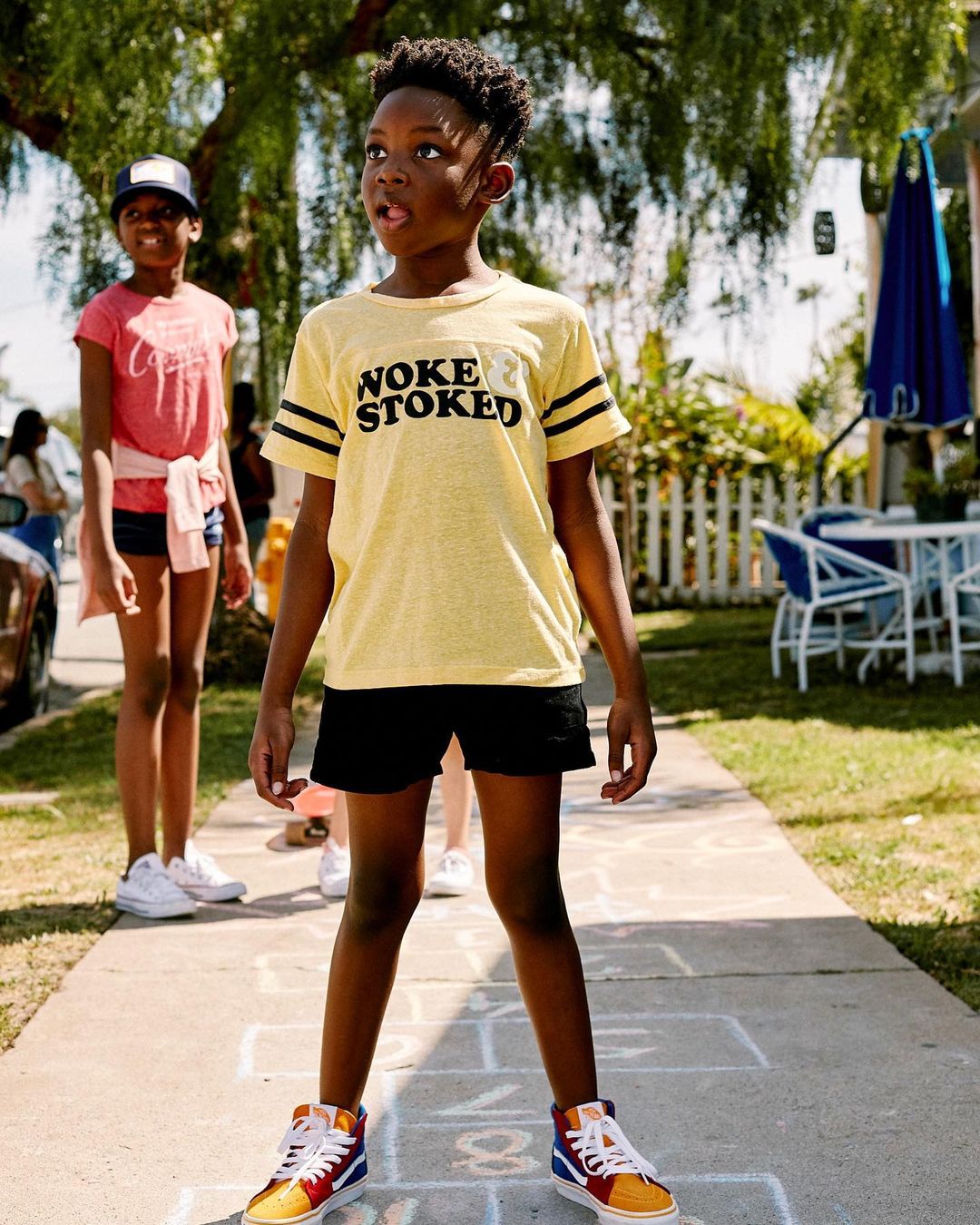 Young boy standing on hopscotch squares outside in a yellow graphic T-shirt, black shorts, and colorful high-top sneakers.