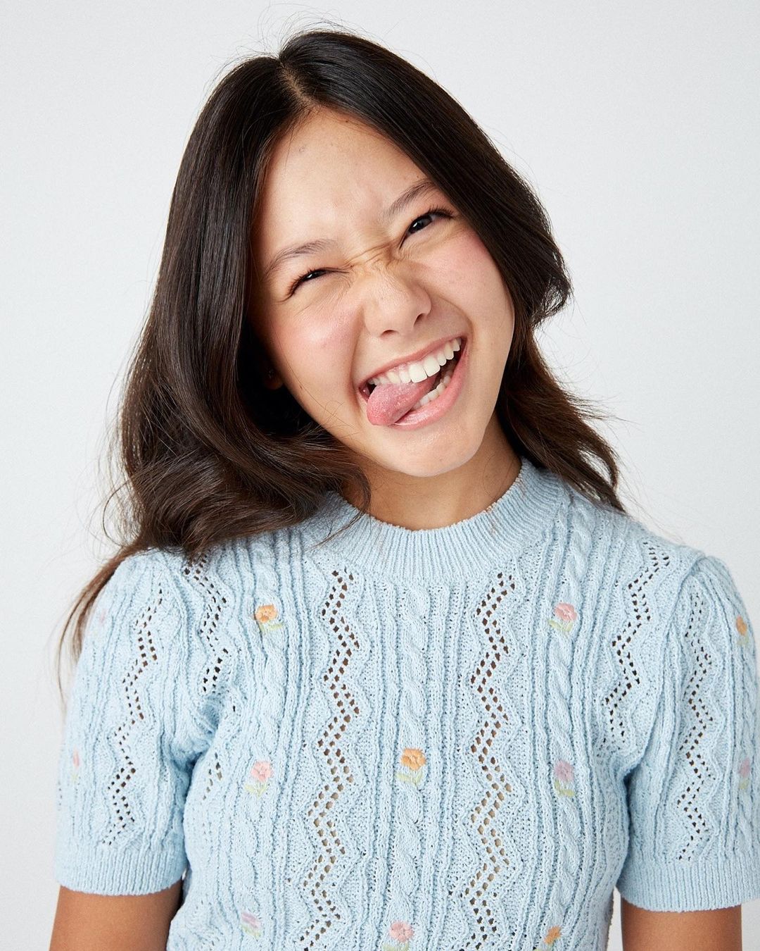 Portrait of an Asian young woman in a light blue sweater sticking her tongue out and squinting with a smile, against a white background.