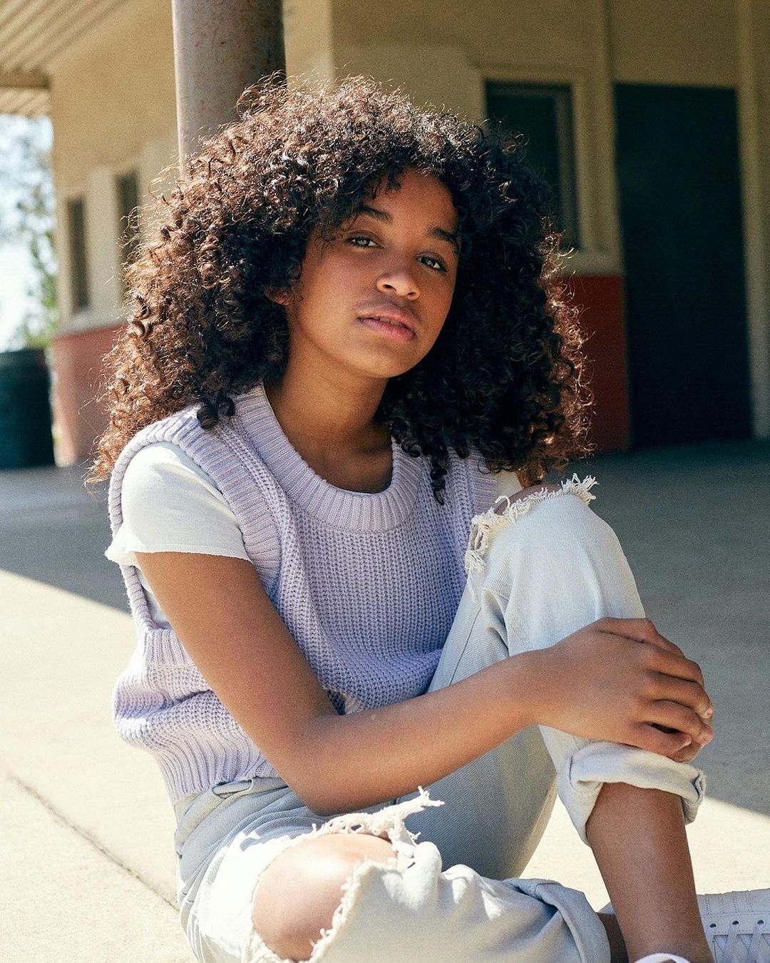 Portrait of a young Black woman with curly hair, wearing a lavender vest and ripped jeans, sitting outdoors with a serious facial expression.
