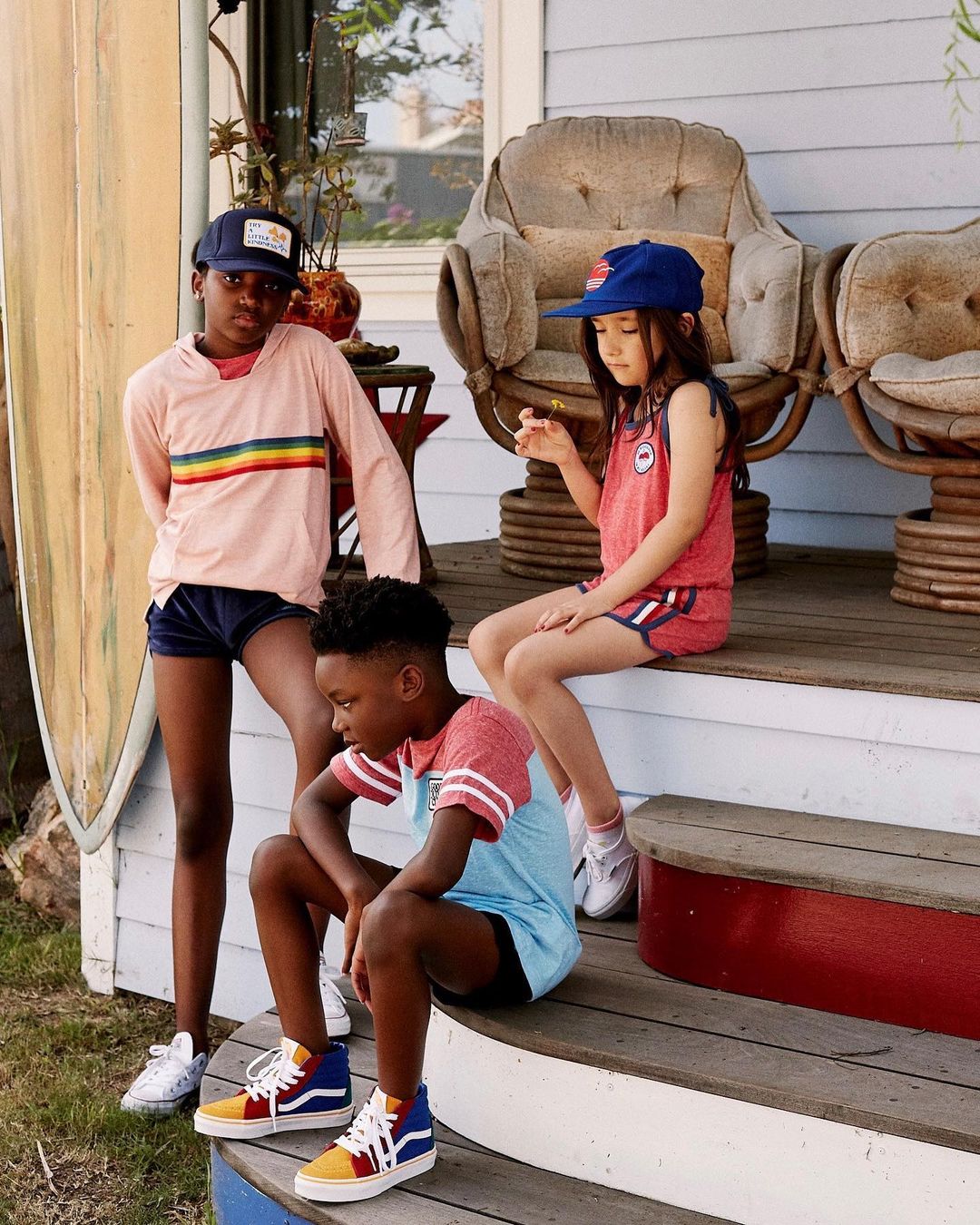Three children of diverse ethnicity wearing colorful clothing and hats are sitting on the steps of a house with a surfboard nearby.