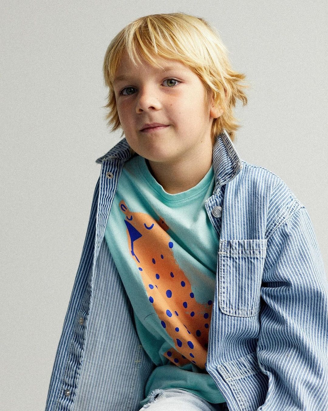 Young boy in a light blue printed t-shirt and vertically striped denim jacket, posing in front of a neutral background.