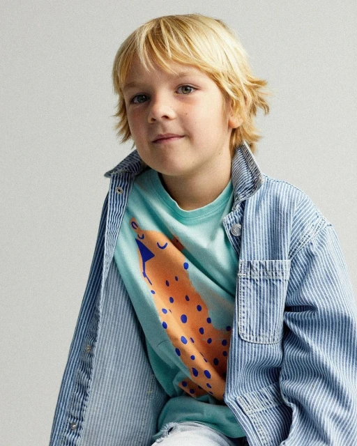 Young boy in a light blue printed t-shirt and vertically striped denim jacket, posing in front of a neutral background.