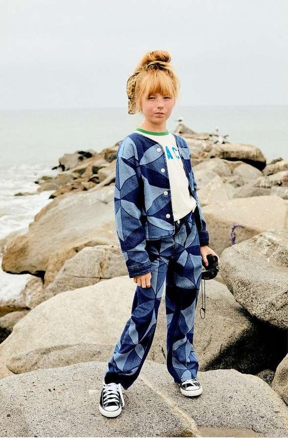Child model poses on rock formation with ocean backdrop, wearing patterned denim jacket and pants, along with black and white sneakers.