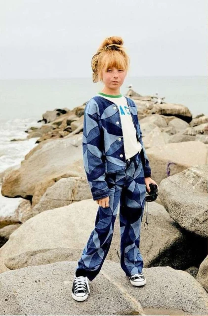 Child model poses on rock formation with ocean backdrop, wearing patterned denim jacket and pants, along with black and white sneakers.