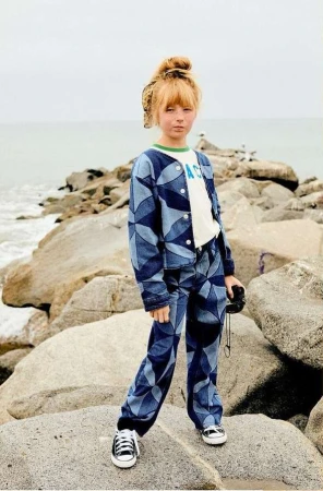 Child model poses on rock formation with ocean backdrop, wearing patterned denim jacket and pants, along with black and white sneakers.