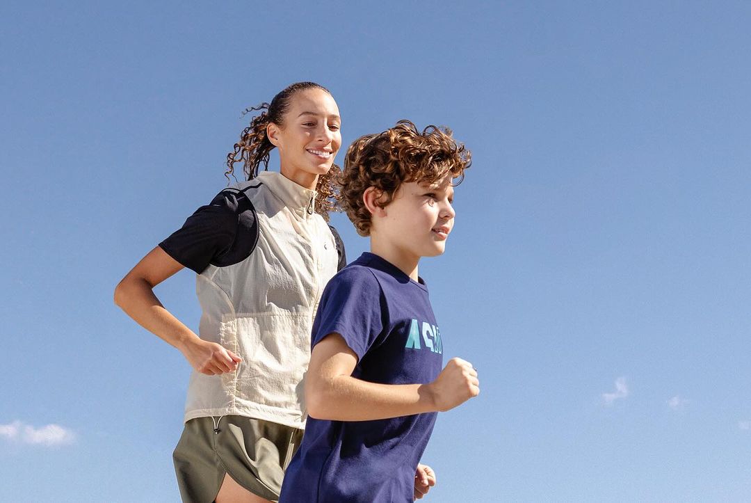 Woman and child run outdoors against clear blue sky. The fair-skinned child wears a blue t-shirt, and the woman wears activewear.