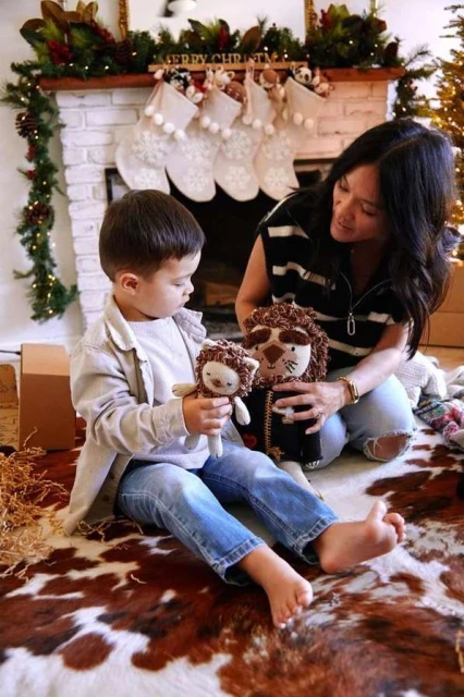 A young child holds stuffed lion dolls with help from an adult female in an indoor setting featuring a fireplace with holiday decorations.