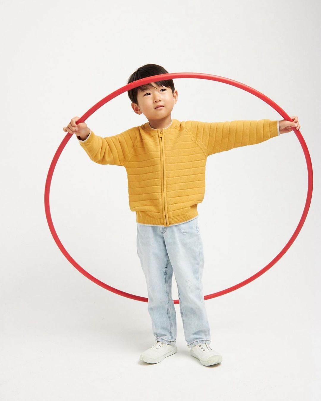 Young Asian child in a yellow sweater holds a red hula hoop while standing in a bright white studio environment.