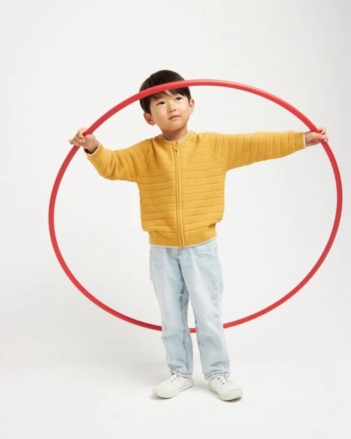 Young Asian child in a yellow sweater holds a red hula hoop while standing in a bright white studio environment.