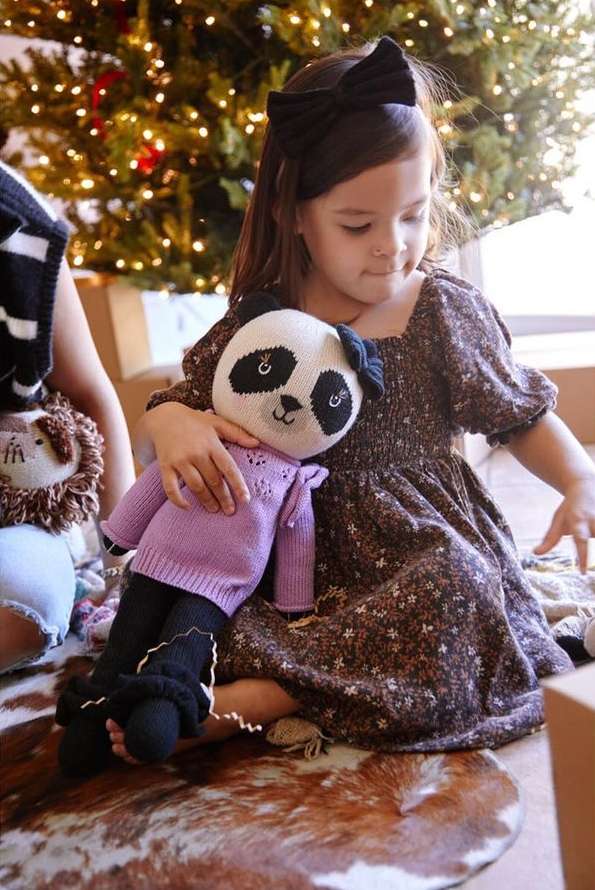Young girl in brown floral dress holds a stuffed panda toy near Christmas tree, with another child and stuffed animal visible.