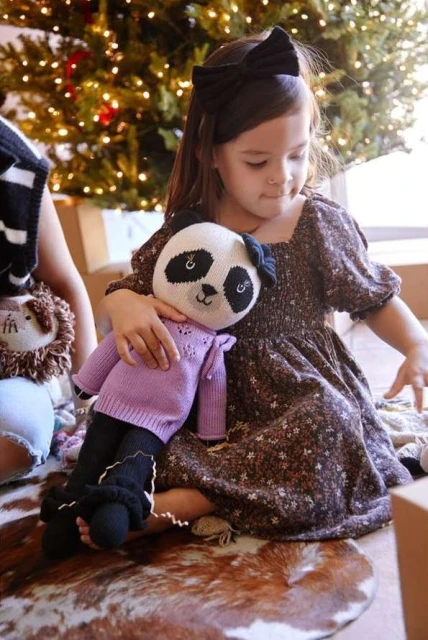 Young girl in brown floral dress holds a stuffed panda toy near Christmas tree, with another child and stuffed animal visible.
