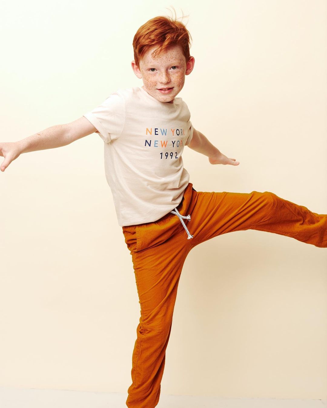 Red-haired child in a studio setting, playfully balancing with arms outstretched, wearing a graphic tee and sweatpants. Bright, cheerful style.