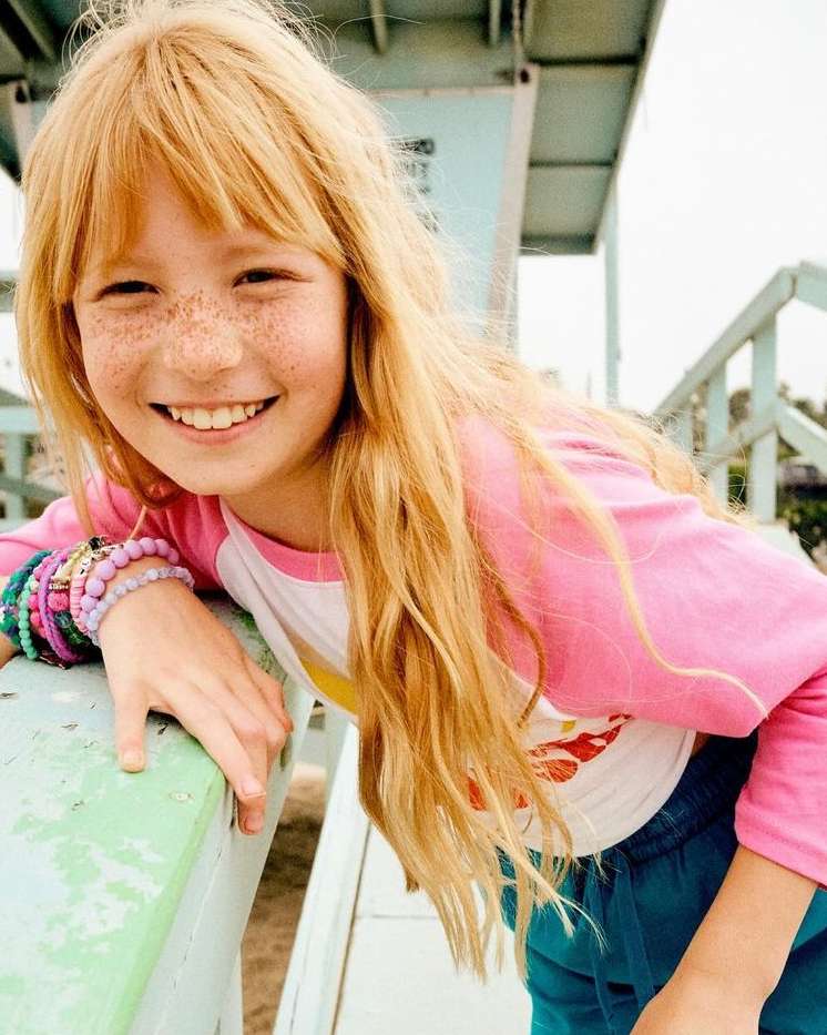 Smiling young girl with freckles and long blonde hair poses in front of light blue lifeguard tower wearing a pink long-sleeved shirt.