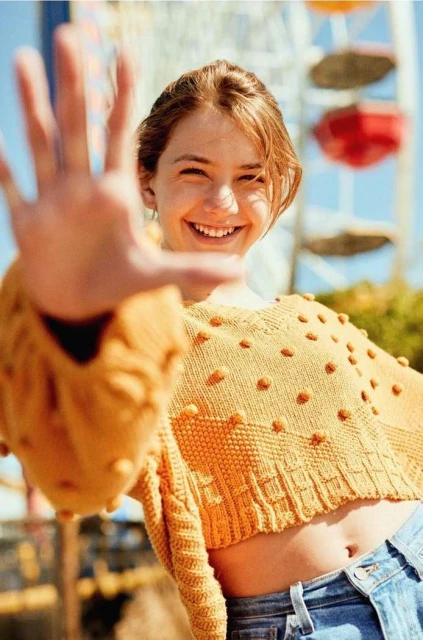 Smiling young woman extends hand toward the camera, wearing an orange sweater and jeans, with a ferris wheel in the background.