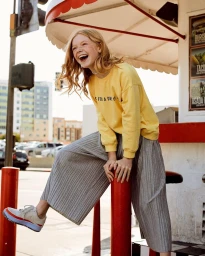 Laughing young red-haired woman wearing a yellow sweatshirt and grey pleated trousers leaning on a red pole outdoors.
