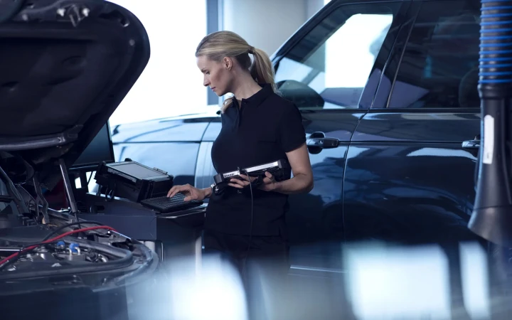 Female mechanic inspecting an engine with diagnostic equipment inside of an auto repair shop, using a handheld device and keyboard.