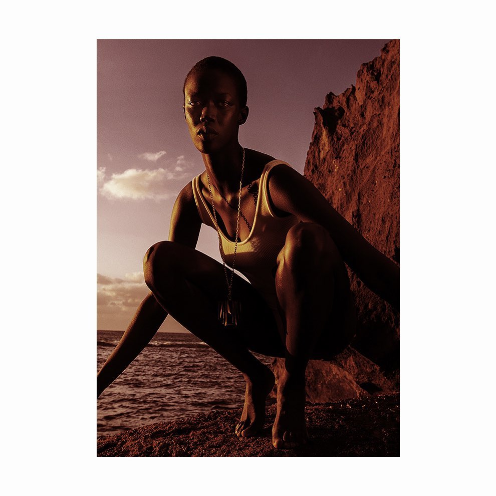 Black woman with a shaved head wearing a tank top and pendant necklace squatting on a rocky shore at sunset