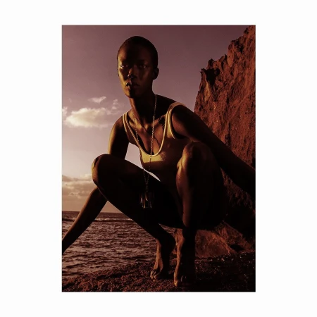 Black woman with a shaved head wearing a tank top and pendant necklace squatting on a rocky shore at sunset