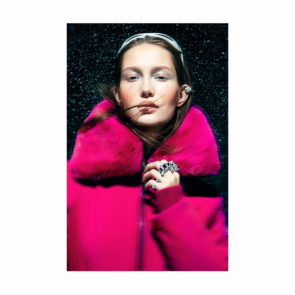 Fashion portrait of a woman wearing a pink coat and jewelry, standing against a black backdrop with water droplets.